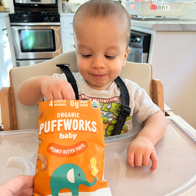Child in a high chair with a package of Organic Puffworks baby snacks in a kitchen setting.