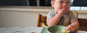 Baby sitting in a high chair eating puffs from a sea green bowl