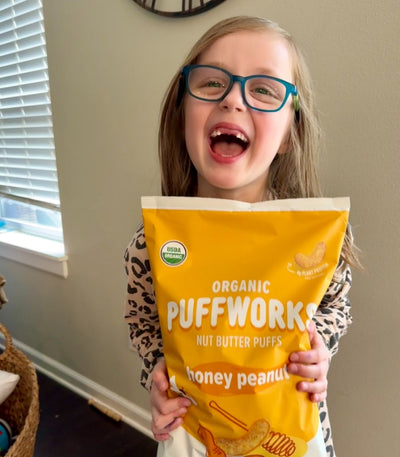Little girl wearing green glasses, smiling and holding a large bag of Puffworks honey peanut nut butter puffs