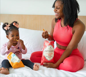 Rai and her daughter smiling and eating Puffworks puffs on a bed with white sheets