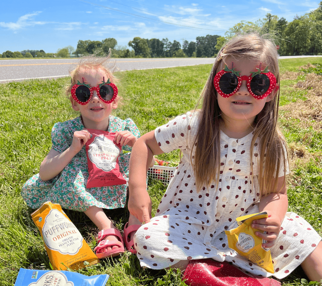 Two girls eating puffworks in a field after strawberry picking, wearing strawberry sunglasses