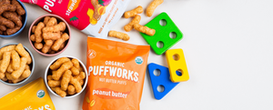 A bag of Organic Puffworks Peanut Butter with colorful kids toys in the back and bowls of puffs